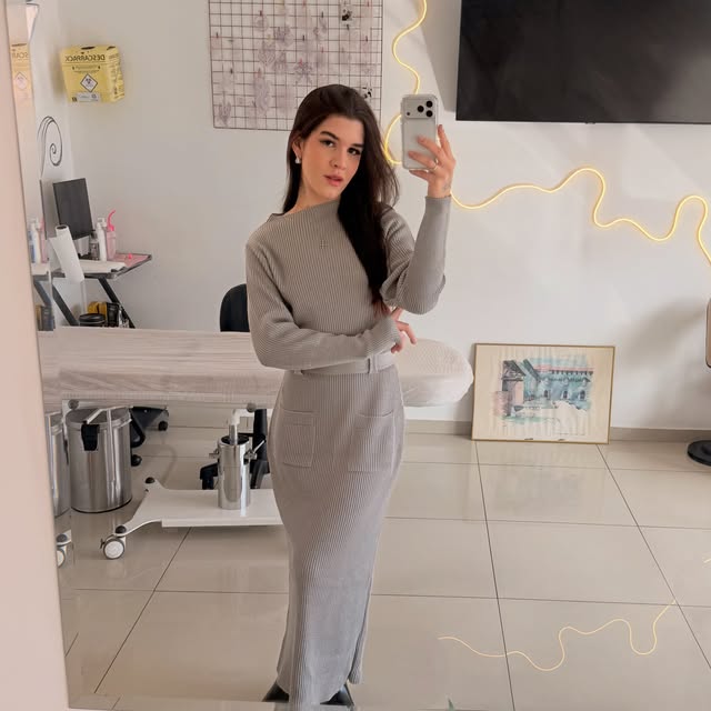 Raquel Damázio no interior do ateliê, com maca, materiais e o neon dourado na parede