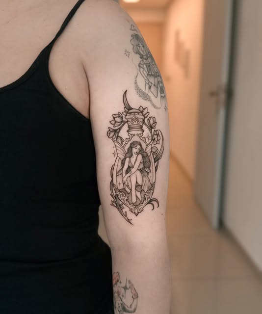 Fada sentada em moldura ornamental com flores, tatuada no braço